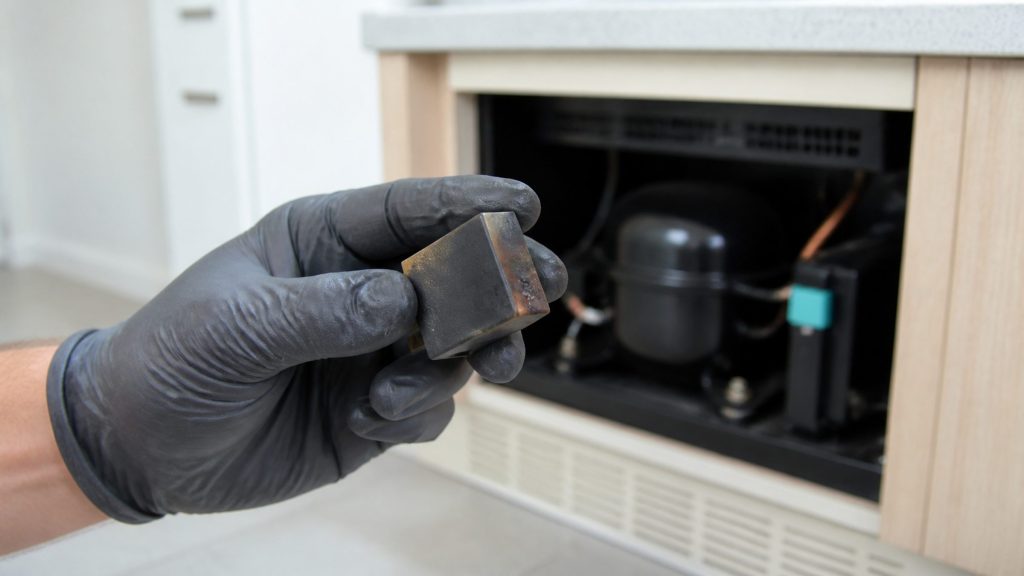 A small black start relay unplugged from the side of a compressor and held in a gloved hand, its plastic body visibly scorched brown at one corner, the machine compartment dark behind it.