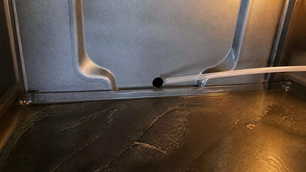 A close view of the small defrost drain hole at the base of the rear wall inside a refrigerator compartment, the shallow channel around it visible, a thin flexible tube approaching it