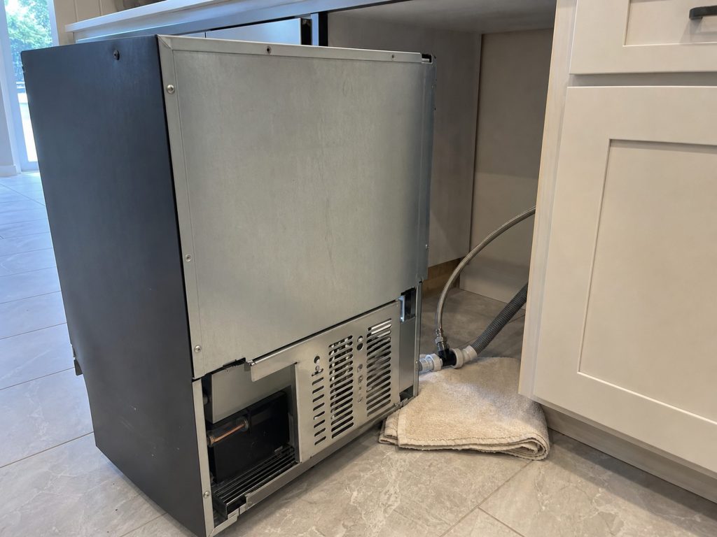 The back of an undercounter ice maker pulled forward on the floor, the water line and the drain pump hose visible behind it, a towel under the joint