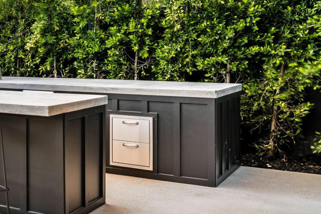 A stainless two-drawer unit built into a dark outdoor kitchen island against a hedge