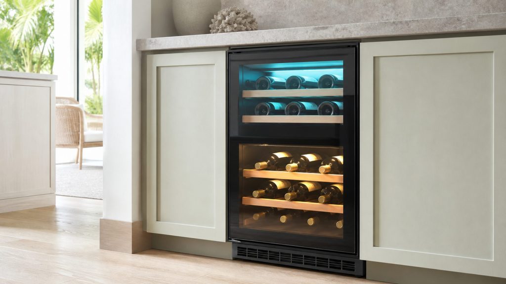 A built-under wine cooler with two temperature zones behind one glass door, the upper zone lit cool and the lower warm, bottles lying on wooden shelves in both.