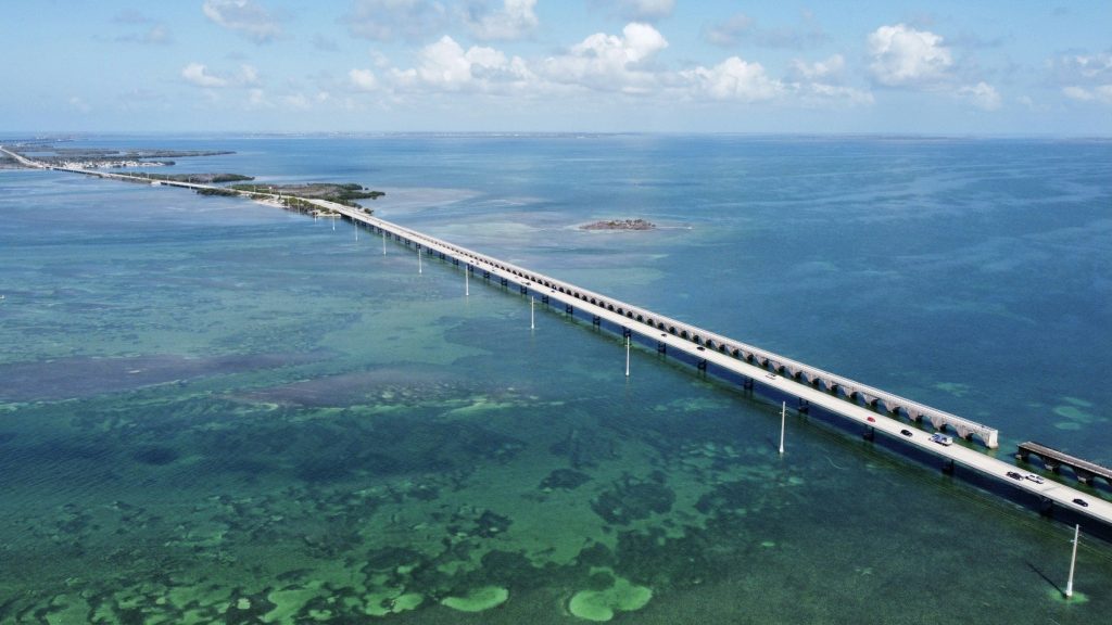 A causeway running low and straight across clear turquoise water under a bright sky, shallows and seagrass visible through the surface either side of it