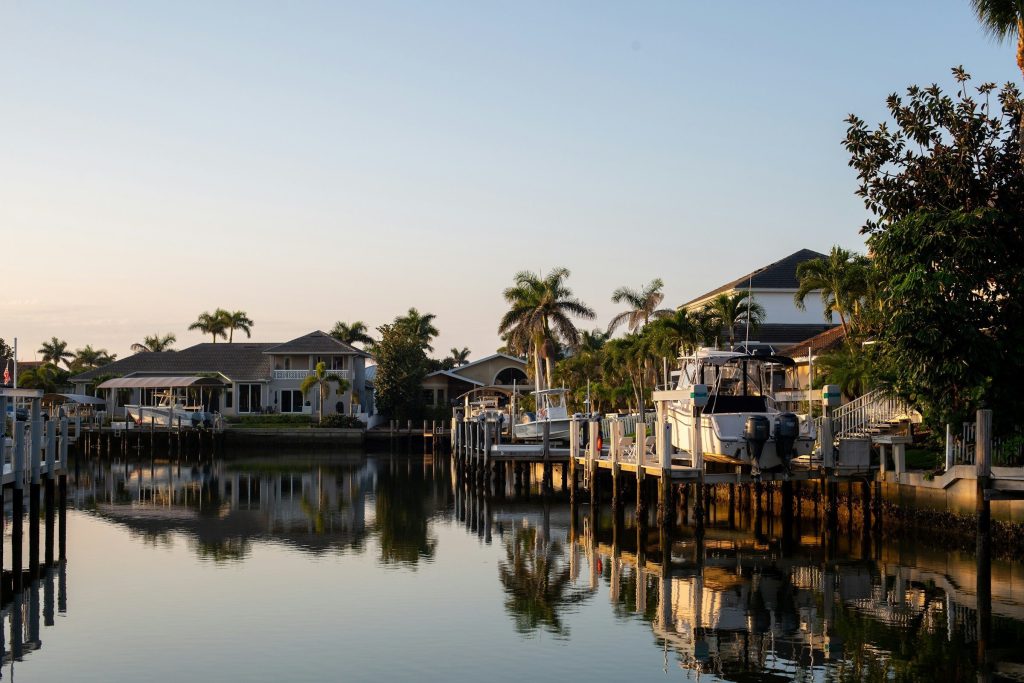 A South Florida canal lined with low waterfront houses and palms in late afternoon light, motor boats moored at private docks, the water still enough to reflect them