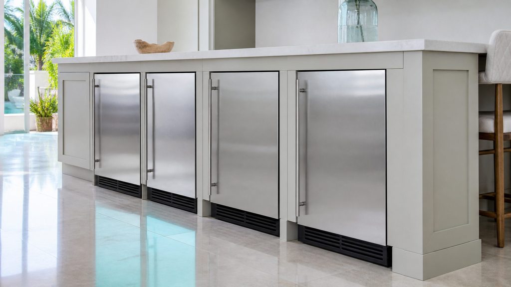 A long run of pale kitchen cabinetry seen low and along its length, four built-under stainless appliance fronts flush in the run beneath a single light stone worktop, a turquoise reflection along the polished floor.