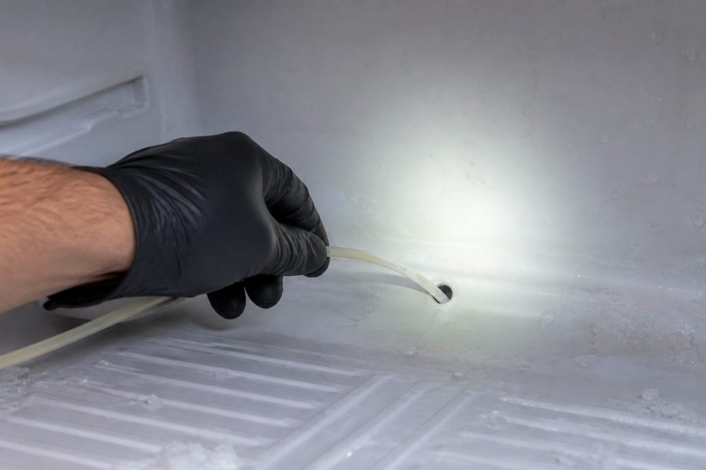 A gloved hand clearing the defrost drain at the base of a freezer compartment with a light shining across the frosted floor