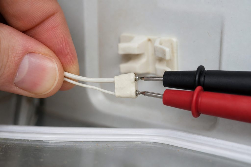 A small thermistor unclipped from its bracket on an interior refrigerator wall and drawn out on its two-wire lead into the cabinet, held between finger and thumb, with a pair of meter probes touching its terminals from the other side of the frame