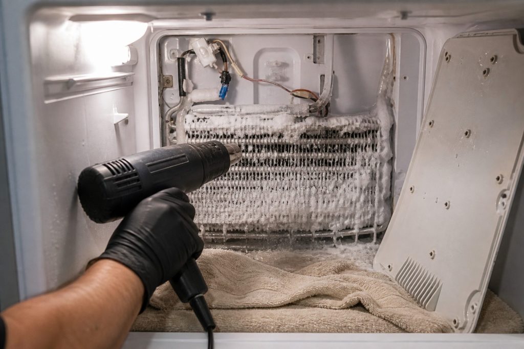 A gloved hand holding a heat gun to a frosted evaporator coil behind the rear panel of a freezer compartment