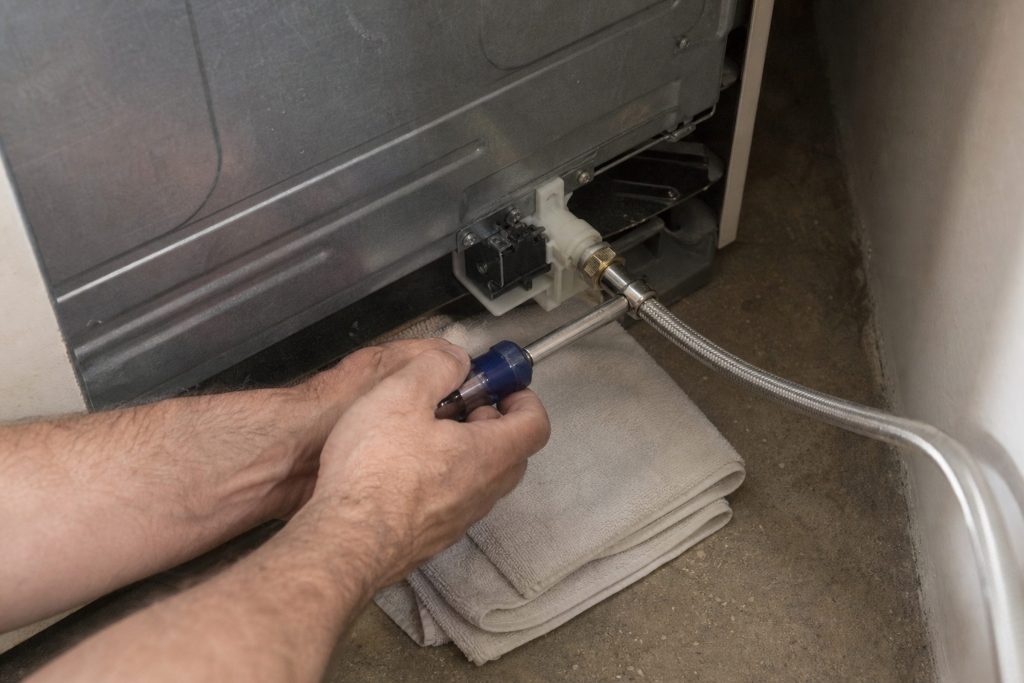 Hands with a nut driver working on the water inlet valve on the lower back of a refrigerator that has been pulled out from the wall, the braided supply line arriving from the right and a folded towel under the fitting
