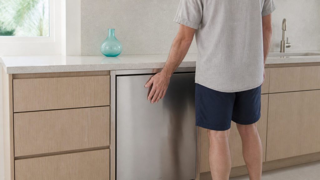 A person standing at a pale kitchen counter with one hand resting on the closed stainless door of a built-under cabinet beneath it, head tilted as if listening, face turned away and out of frame, the room otherwise still.