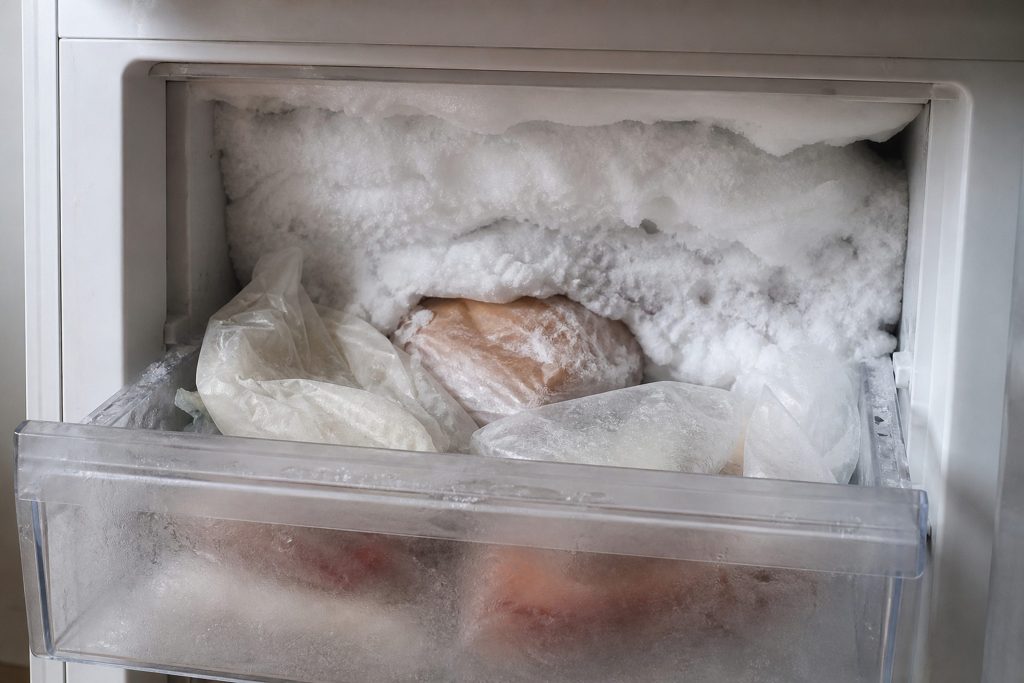The back wall of a freezer compartment carrying a thick white ridge of frost, packages half buried in it, the drawer pulled forward