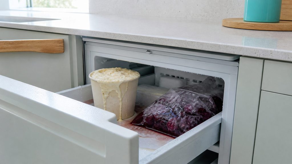 An open built-under freezer drawer where the food has plainly thawed — a tub of ice cream slumped soft at the edge, a bag of berries wet and collapsed, condensation beading on the packaging, photographed close in cold light.