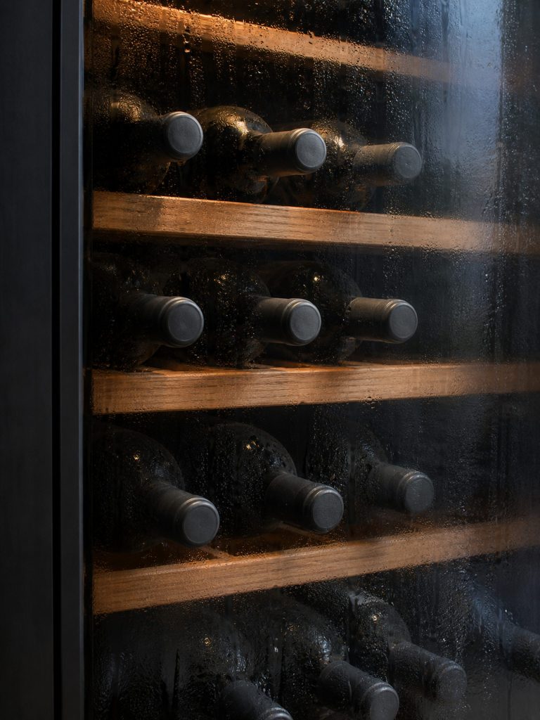 Wine bottles lying on wooden shelves behind smoked glass, the inside of the glass fogged with condensation